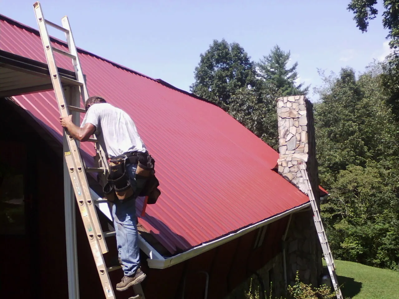 Roofer on a ladder beside a red metal roof and stone chimney — Soffit & Fascia Repair by Roofing Brothers in Lake Huntington